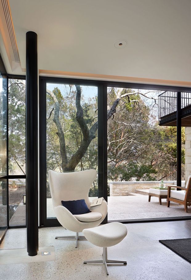 A bright modern living room with full-height black-framed glazing overlooking mature trees and a paved outdoor terrace. The room features a sculptural white lounge chair with matching ottoman, a polished concrete floor, a slim black cylindrical fireplace or structural element, and an outdoor balcony or terrace visible beyond the glass.
