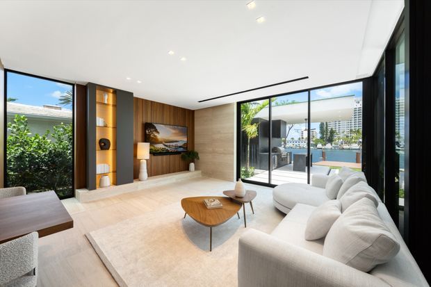 Bright contemporary living room with floor-to-ceiling glazing, a large pale sectional sofa, light rug, sculptural wood coffee table, and a timber-and-dark-panel built-in media wall. The room opens visually to lush planting and a waterfront setting beyond the glass.