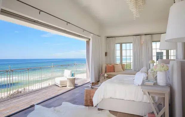 Bright coastal bedroom with a large open glazed wall overlooking the ocean and beach. The room features a white upholstered bed with layered white bedding, pale upholstered seating, sheer curtains, light timber side tables, a woven stool, and a glass pendant chandelier. A spacious timber deck with metal balustrading extends outside, creating a relaxed beachfront setting.