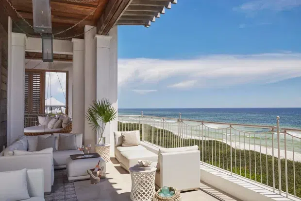 A bright, refined coastal outdoor lounge terrace overlooking a sandy beach and ocean. The covered terrace features white upholstered sofas and armchairs, a low coffee table, decorative side tables, potted palms, timber ceiling detailing, large white columns, and a white balustrade. Shutters and additional seating are visible further inside, while the open side frames expansive sea views.