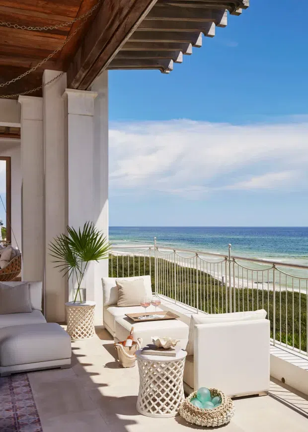 A bright coastal outdoor lounge terrace overlooking the ocean. The covered terrace has white upholstered seating, a low coffee table, decorative lattice side tables, potted palm, patterned tile flooring, white balustrades, and a substantial timber roof with exposed rafters and decorative chain details. The beach and sea are visible beyond the railing.