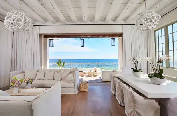 Bright coastal living and dining space opening directly onto an ocean view. The room features white-painted exposed ceiling boards and beams, full-height white curtains, large multi-pane windows, light timber flooring, a cream sectional sofa, and a long white dining table with slipcovered chairs. Woven and organic decorative elements, orchids, and sculptural pendant lights create a relaxed, refined beach-house atmosphere.