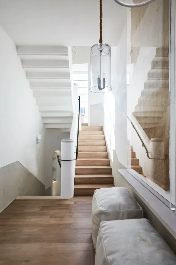 Bright contemporary stair hall with a central timber staircase, an adjacent stair flight reflected in a large wall mirror, white walls and balustrades, warm timber flooring, and a suspended cylindrical glass pendant light. A long upholstered bench sits along the right side beneath the mirror, while a lower stairwell and sculptural white railing element add depth.