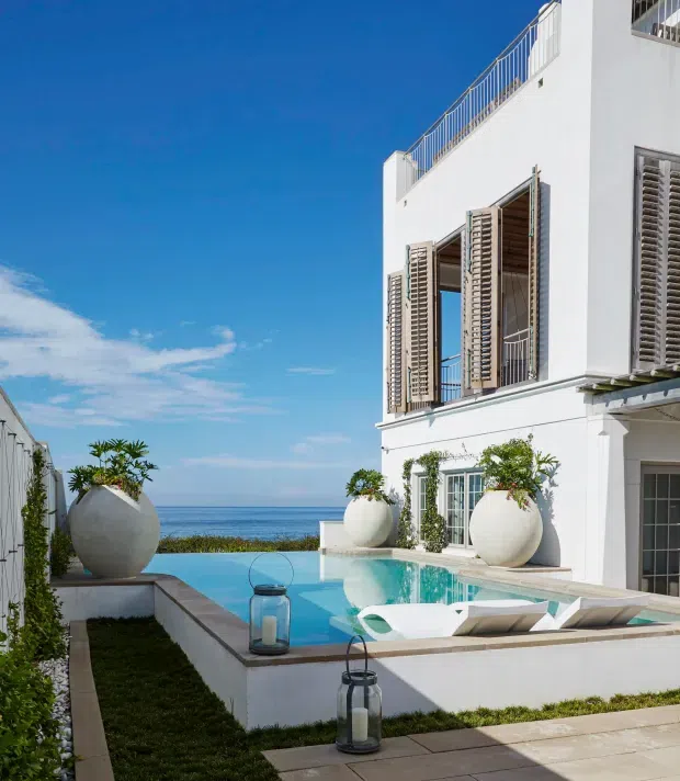 Bright coastal exterior of a white multi-storey residence beside the sea, featuring a compact turquoise swimming pool, pale stone paving, large spherical planters, and tall timber louvred shutters opening onto balconies and glazed doors. A rooftop terrace with metal balustrading is visible, with strong blue sky and ocean beyond.