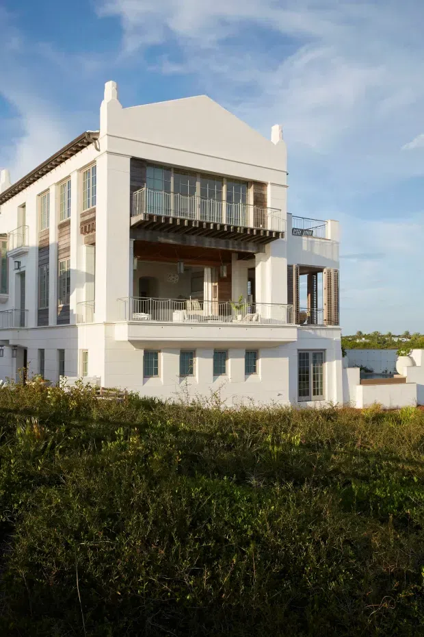 Exterior photograph of a large white multi-storey residence with a formal, symmetrical facade, tall multi-pane windows, projecting balconies, open terraces, metal balustrades, and chimney-like parapet details. The building is set behind landscaped vegetation under a bright blue sky.