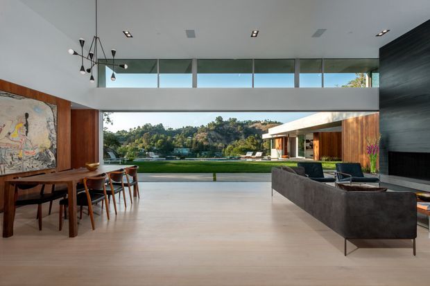 A spacious, sophisticated open-plan living and dining area with floor-to-ceiling glazing overlooking a landscaped lawn and wooded waterfront setting. The room features pale timber flooring, dark timber wall panelling and joinery, a long timber dining table with sculptural chairs, a contemporary grey sofa, a dramatic black textured fireplace wall, and a double-height ceiling with suspended decorative lighting.