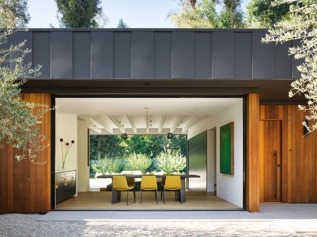 Front exterior view of a contemporary single-storey home with a wide, fully opened glazed dining pavilion or garage-like opening. The facade combines dark vertical metal cladding, warm timber wall cladding and a timber entry door. Inside, a dining table with yellow chairs sits beneath an exposed white-beam ceiling, with extensive glazing opening toward landscaped greenery.