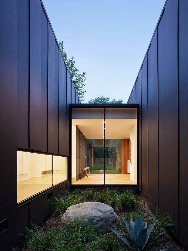 A dramatic exterior view of a contemporary dark-clad building forming a narrow landscaped courtyard. Tall flat wall planes frame a brightly illuminated glazed entry or living space, with warm timber visible inside. A large natural boulder, ornamental grasses, and sculptural planting occupy the foreground, while the symmetrical composition emphasizes the recessed glass pavilion and vertical facade walls.