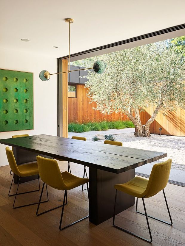 A contemporary dining room with a large dark timber dining table surrounded by mustard upholstered chairs. Full-height glazed sliding doors open directly to a landscaped courtyard containing an olive tree, gravel, planting and timber fencing. The room has pale walls, timber flooring, a linear ceiling vent or track, and a sculptural brass-and-glass pendant light. A green textured artwork is mounted on the wall.