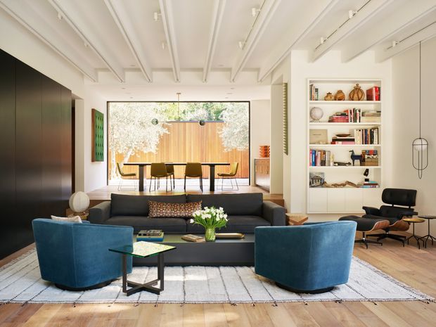 Bright, refined living room with a contemporary open-plan layout. The space features a large dark sofa, two deep blue lounge chairs, a low coffee table, a patterned area rug, timber flooring, and extensive built-in shelving. A dining area is visible beyond the living zone through full-height glazing, while exposed white ceiling beams and track lighting add architectural character. The neutral white, timber, charcoal, and blue palette creates a warm yet clean atmosphere.