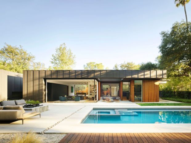 A low, single-storey modern residence viewed from the pool terrace. The house has a broad flat roof, dark metal-clad facade, extensive floor-to-ceiling glazing, warm timber doors, and an open indoor-outdoor living connection. A rectangular swimming pool, pale paved terrace, and timber decking occupy the foreground, with mature landscaping around the property.