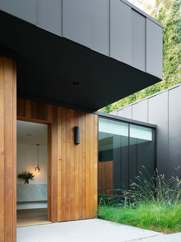 Contemporary residential entrance viewed from an exterior courtyard or walkway. The facade combines vertical warm timber cladding with dark charcoal metal panels, deep cantilevered roof planes, and large glazed openings. An open timber door reveals a refined interior with a stone-textured wall, timber flooring, and pendant lighting. Ornamental grasses and dense greenery soften the dark, geometric architecture.