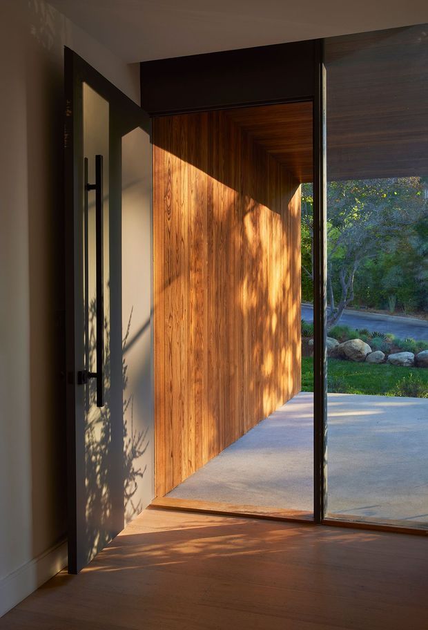 A warmly lit modern entryway viewed from inside, featuring a tall pivot door with a dark metal frame and long vertical handle, a richly grained timber-clad entry wall and soffit, timber flooring, and adjacent full-height glazing opening toward a landscaped exterior. Strong sunlight creates dramatic shadows across the door, wall, and floor.