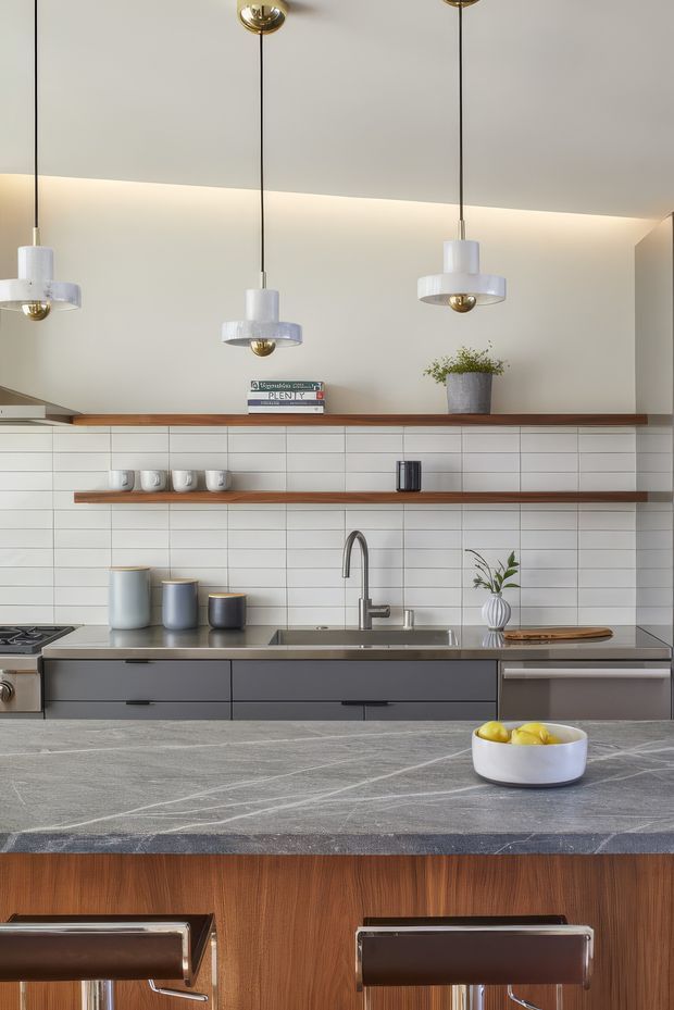A contemporary kitchen viewed straight on, featuring dark grey lower cabinetry, a stainless-steel sink and appliances, white horizontal subway-tile backsplash, open timber shelves, a large grey stone island with timber-clad base, and suspended glass-and-metal pendant lights. Neutral tones are balanced by warm wood accents and a bowl of lemons on the island.