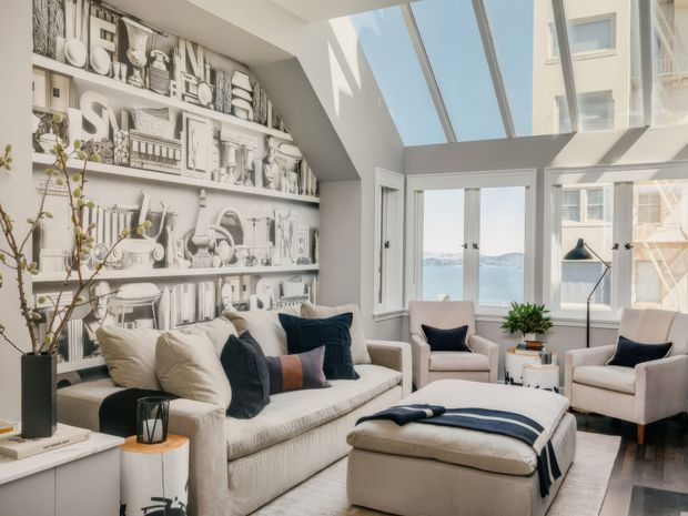 Bright, refined living room with a large cream sectional sofa, matching upholstered ottoman, two armchairs, and a small side table. A dramatic monochrome illustrated feature wall with built-in shelving fills the left side, while tall windows and roof glazing bring in abundant daylight and offer a water view. The space has a light neutral palette with navy and muted accent cushions, contemporary furnishings, and a clean, inviting atmosphere.