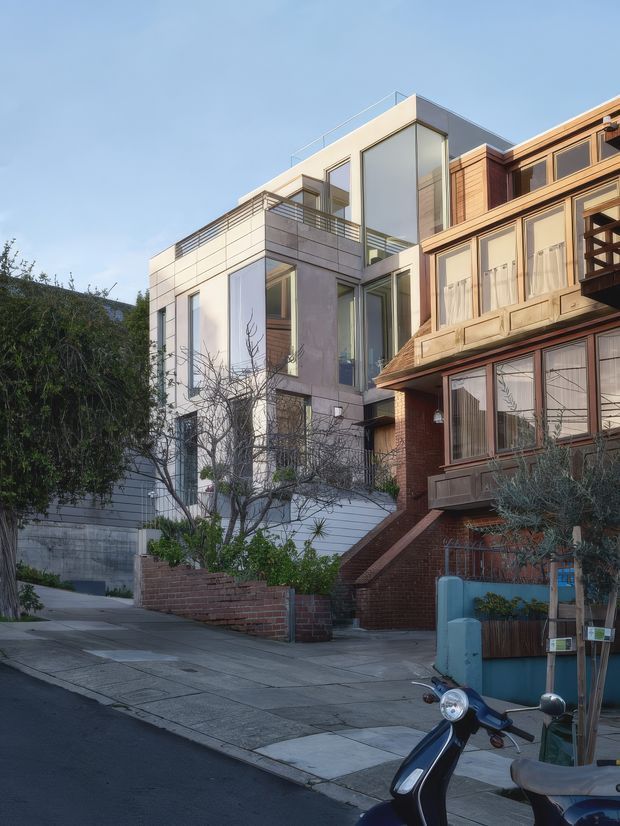 Street-level exterior view of a contemporary multi-storey urban residence, showing a white rendered and glazed modern extension beside an older brick-clad dwelling. The newer building has strong rectangular volumes, extensive floor-to-ceiling glazing, a rooftop terrace with glass balustrade, and horizontal metal railings. The adjoining older facade features timber-framed windows, projecting bay elements, brickwork, and a pitched roof edge. A paved sidewalk, small front gardens, retaining walls, and a scooter occupy the foreground.