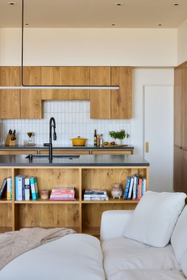 A warm, contemporary open-plan living room and kitchen. The foreground features a large white upholstered sectional sofa, while a timber open-shelving room divider separates it from a compact kitchen. The kitchen has extensive natural timber cabinetry, a dark benchtop and island, white vertical-grid tiled splashback, black tapware, and a linear suspended black pendant light. Built-in timber cabinetry continues along the right wall, with a concealed white door adding to the streamlined composition.