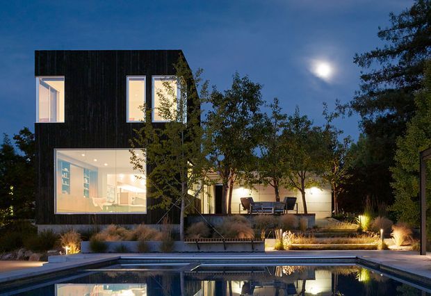 Twilight exterior view of a contemporary two-storey residence with a dark vertically clad upper volume, large illuminated windows, and a brightly glazed ground-floor living area. The home opens onto a landscaped terrace with ornamental grasses, warm architectural lighting, and a rectangular swimming pool in the foreground.