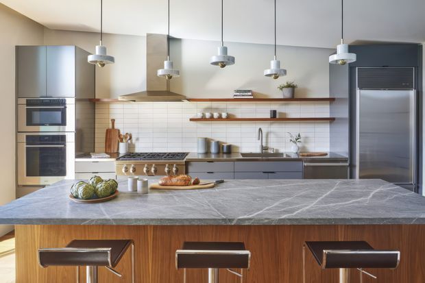 A spacious contemporary kitchen viewed from behind a large island. The room features dark grey fixed cabinetry, integrated stainless-steel appliances, a white horizontal subway-tile splashback, open timber shelves, and a substantial grey veined stone island with a warm timber-clad base. Three black leather bar stools sit at the island, while multiple suspended pendant lights hang from the ceiling. The composition is clean, functional, and softly modern with warm timber accents.