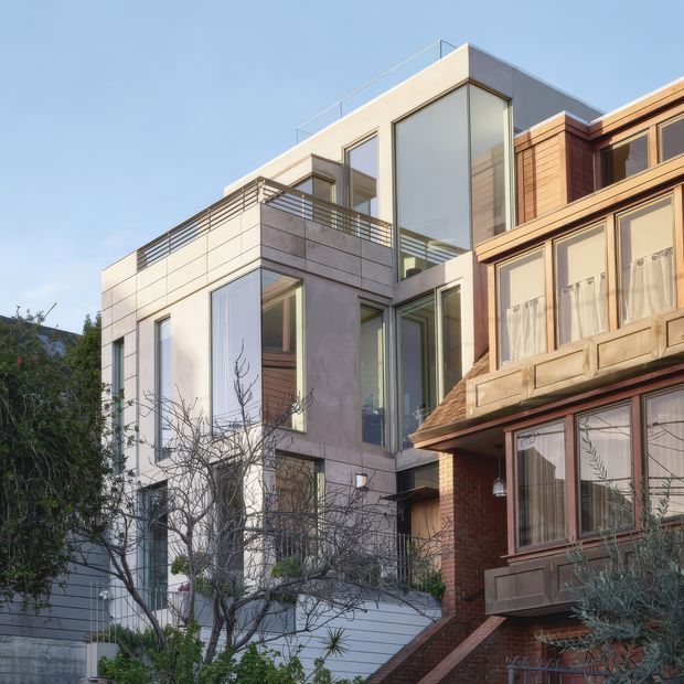 Exterior view of a contemporary multi-storey urban residence adjoining an older traditional brick dwelling. The newer building has a flat roof, large floor-to-ceiling windows, pale concrete or rendered wall panels, a rooftop terrace with slim metal balustrades, and warm timber-toned cladding. The surrounding mature residential streetscape is visible but secondary.