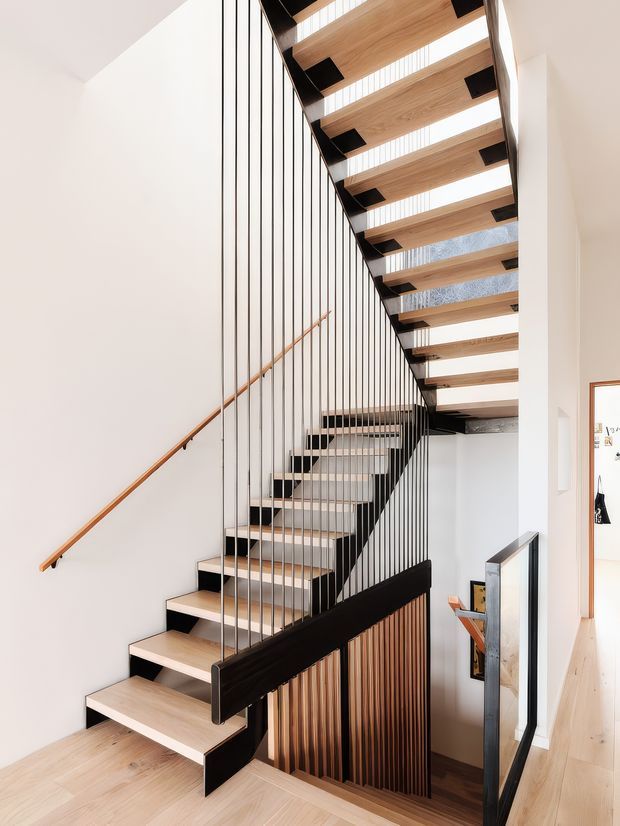 A bright, contemporary interior stairway with open-riser timber stairs, black steel stringers and slim vertical balustrade rods. The staircase rises beneath a timber soffit with integrated skylight-like glazed strips, while a timber handrail and slatted timber screening add warmth against white walls and pale timber flooring.