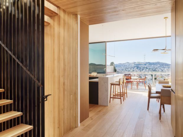 Warm contemporary interior looking from a stair and entry zone into an open-plan kitchen and dining area. Vertical timber wall panelling and a slatted timber ceiling frame pale timber flooring, while a dark open-riser stair and black metal screen create contrast. The kitchen has a stone island, timber cabinetry, bar stools and a dining setting, with expansive glazing opening views toward a densely built hillside landscape.