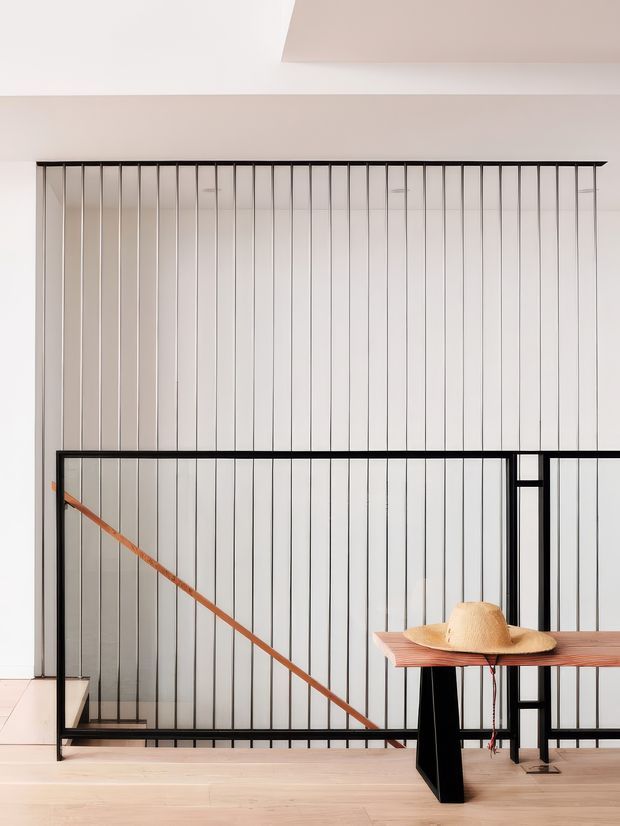 Bright minimalist interior showing a stairwell or landing enclosed by a black metal balustrade with closely spaced vertical rods and a warm timber handrail. Pale timber flooring and a simple timber console or table with a straw hat occupy the foreground. The space has a high ceiling and an airy, refined architectural character.
