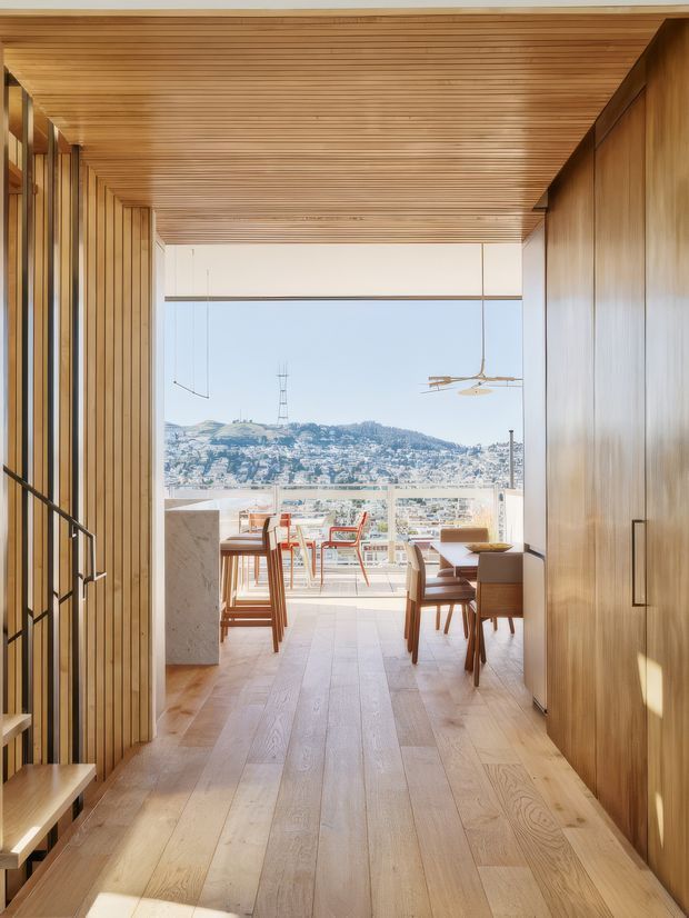 A warm, wood-lined interior passage opens onto a bright dining and living area with expansive glazing and a balcony or terrace beyond. The room features a timber slatted ceiling, vertical timber wall panelling, wide light-oak flooring, built-in timber cabinetry, a stone-clad island or counter, a dining table with chairs, and a panoramic view of a densely built hillside city. A sculptural ceiling fan hangs in the glazed space, while a stair and open shelving are visible along the left side.