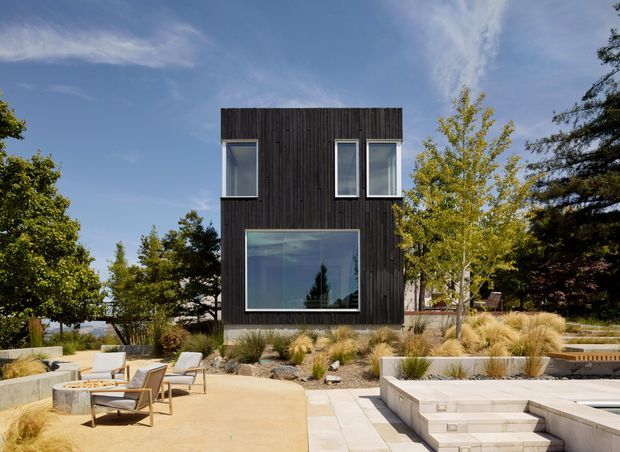 A contemporary two-storey dark-clad residential building viewed from a landscaped outdoor entertaining area. The compact rectangular facade features vertical dark timber cladding, large minimalist windows, and a broad ground-floor glazed opening. In the foreground are pale paved terraces, low rendered retaining walls, built-in outdoor seating or steps, and ornamental grasses, with mature trees surrounding the site.