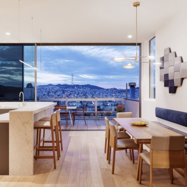 A bright, contemporary open-plan kitchen and dining area with expansive floor-to-ceiling glazing overlooking a densely settled hillside and city lights. The room features a pale marble kitchen island with integrated sink, light timber flooring, a timber dining table with upholstered dining chairs, bar stools, a built-in blue banquette, and sculptural modern pendant lighting. A balcony or glazed outdoor edge is visible beyond the dining area.