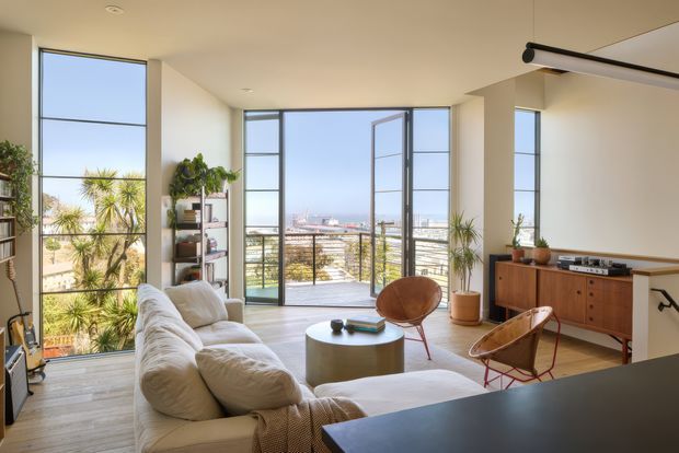 Bright contemporary living room with floor-to-ceiling steel-framed glazing and an open balcony overlooking a coastal urban landscape. The room includes a large cream sectional sofa, round coffee table, mid-century-inspired timber sideboard, leather sling chair, indoor plants, and a dark kitchen island edge in the foreground. Natural light, pale finishes, and expansive windows create an airy, relaxed atmosphere.