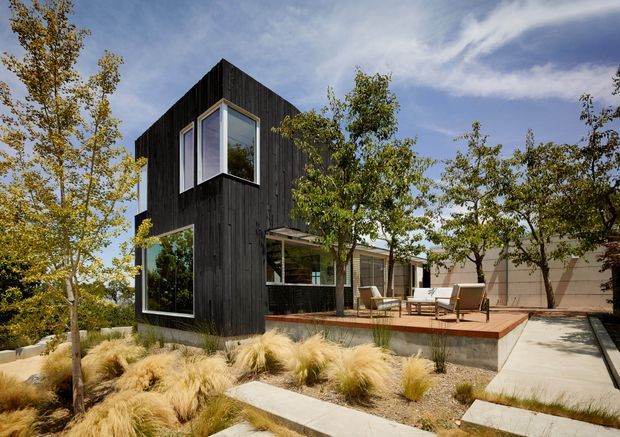 Contemporary compact house with a dramatic black vertical-clad upper volume projecting over a lower glazed living area. The home sits in a landscaped suburban garden with ornamental grasses, mature trees, concrete stepping paths, and a raised timber-toned deck furnished as an outdoor seating area. Large corner windows and a steeply pitched asymmetrical roofline create a strong modern architectural character.