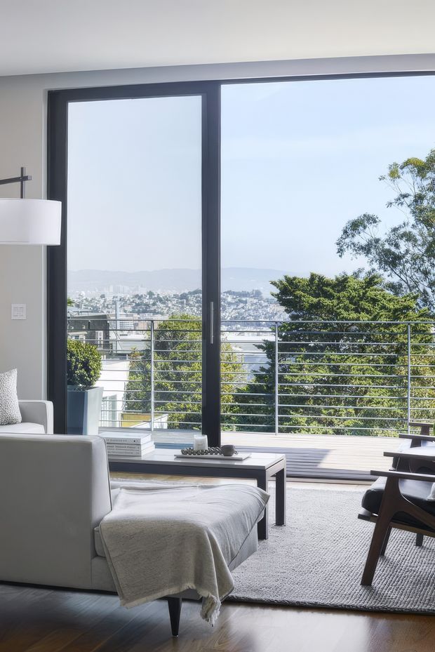 Bright contemporary living room with floor-to-ceiling dark-framed glazing opening onto a timber deck and horizontal metal balustrade. The room contains a pale upholstered armchair with a draped throw, a low black coffee table, textured light rug, and dark timber furniture. A leafy suburban hillside cityscape is visible beyond the terrace.