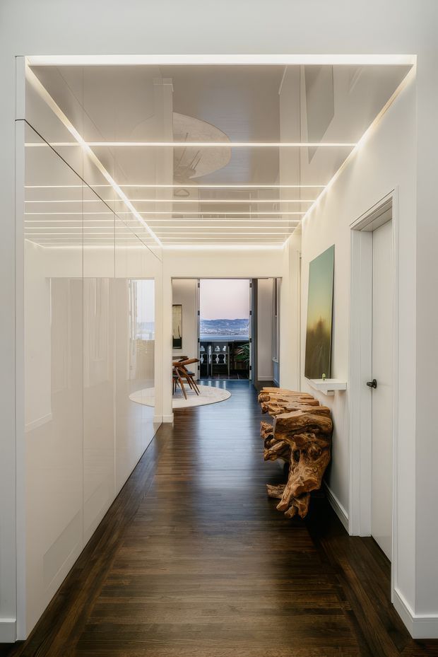 A long, refined interior hallway with dark timber flooring, white walls and built-in cabinetry, a mirrored wall to the left, and a linear illuminated ceiling grid. A sculptural raw timber console runs along the right wall, leading toward an open dining or living area with views beyond.