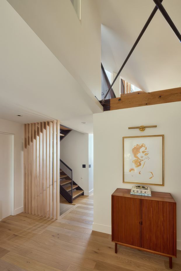 Interior view of a bright, refined entry hall with pale timber flooring, white walls, and a sculptural open-riser staircase. Vertical timber screening partially separates the stair and circulation area. A timber credenza with fluted detailing, framed artwork, and a brass wall light forms a styled focal point. Exposed timber beams and dark metal tension bracing are visible above, suggesting a high-level void or open stair volume.