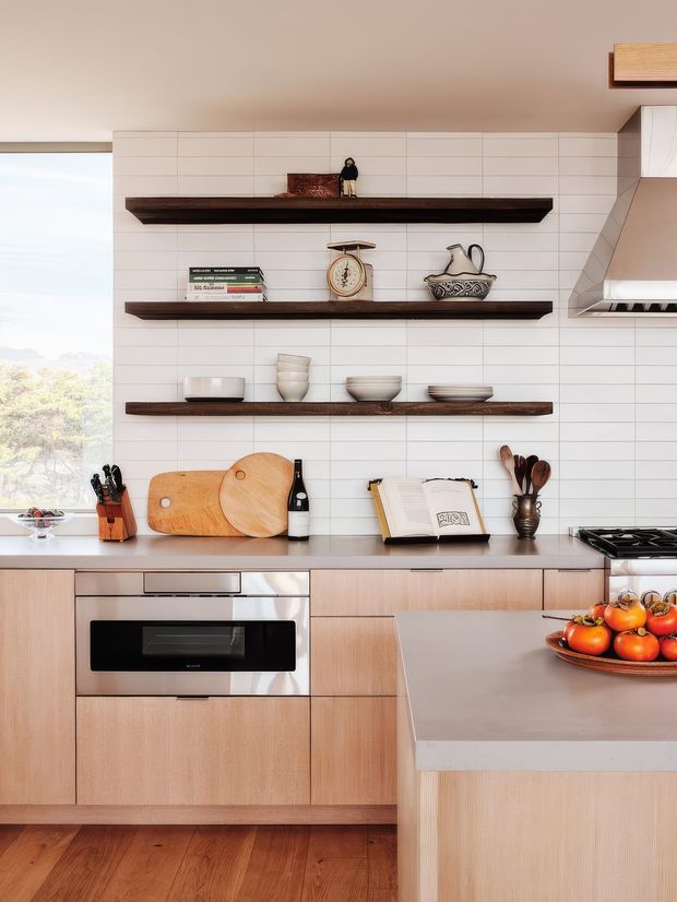 Bright contemporary kitchen with light timber cabinetry, a white horizontal tile backsplash, dark floating timber shelves, stainless-steel appliances, and a substantial island with a pale benchtop. Open shelving displays ceramics, books and kitchenware, while a large window brings in natural light and views of greenery.