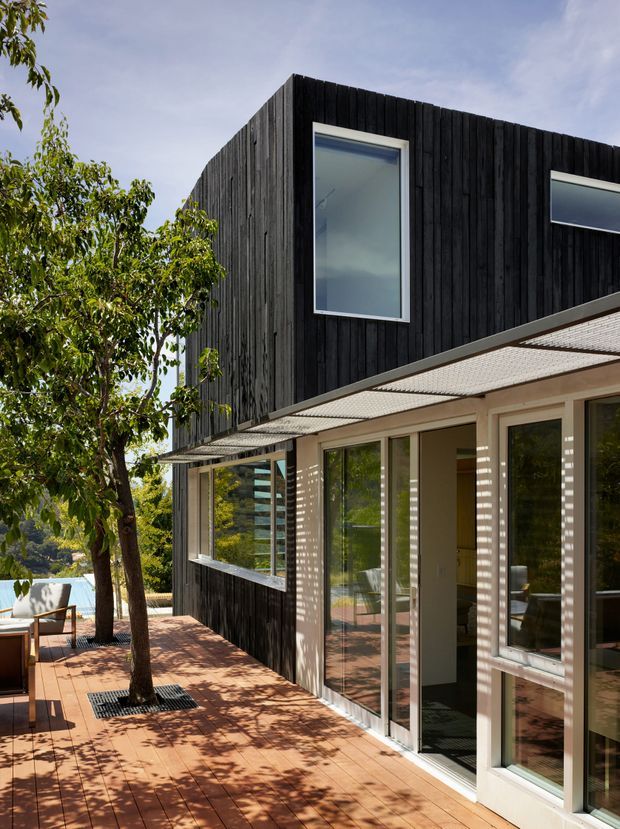 Exterior architectural photograph of a modern two-storey house with a dark vertically clad upper volume, pale lower-level walls, expansive glazed doors and windows, and a broad perforated shade canopy. A timber deck with integrated tree planting runs alongside the facade, with outdoor seating visible at the edge.