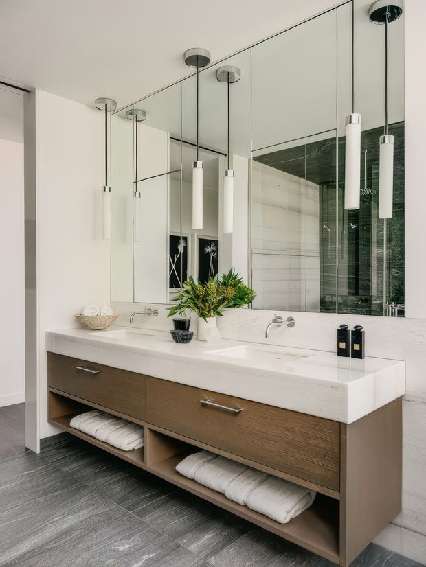 A contemporary double vanity bathroom with a floating timber-look vanity, white stone benchtop, integrated basins, wall-mounted chrome tapware, large mirrored wall panels, and cylindrical pendant lights. Dark grey floor tiles and a green-toned tiled shower enclosure add contrast, while open vanity shelving stores folded towels.