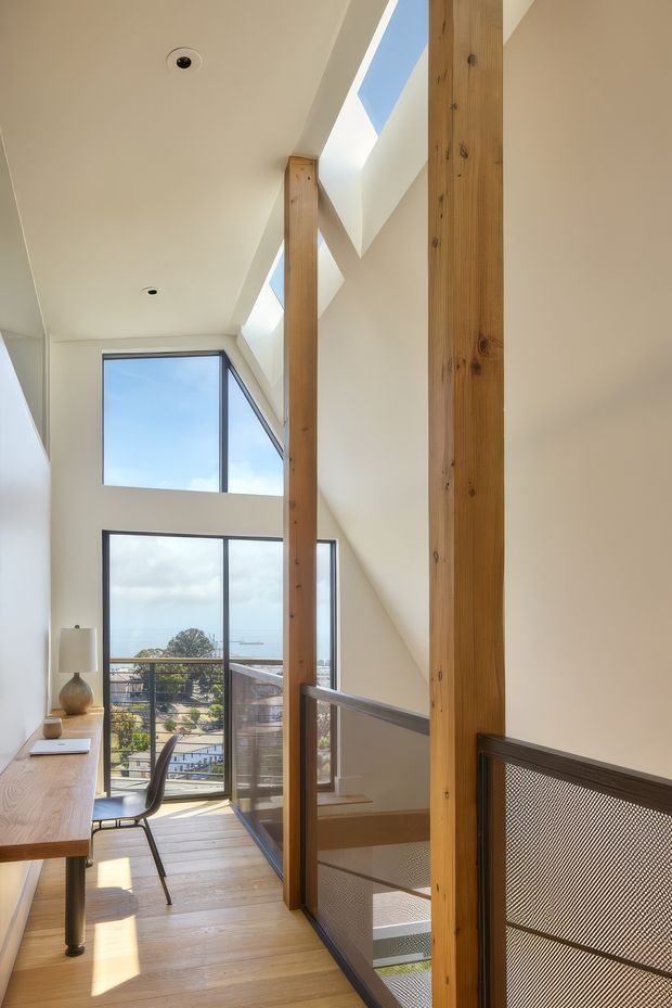 Bright, narrow multi-level interior corridor or landing with a compact timber-floored workspace along the left wall. Full-height glazing and angular skylights bring in abundant daylight and reveal a distant coastal or urban view. Exposed vertical timber posts and beams frame the space, while a dark metal mesh balustrade guards the stair opening on the right. The interior has a clean contemporary architectural character with natural timber and white plastered surfaces.