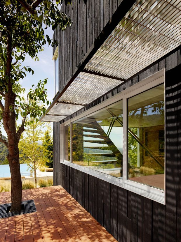 Exterior view of a contemporary building with dark vertically charred-looking timber cladding, extensive horizontal glazing, and projecting translucent slatted sunshades. A timber deck and mature trees frame the facade, while an angular interior stair is visible through the windows.