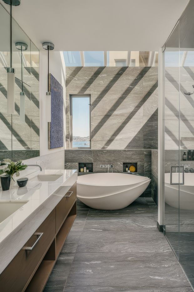 A bright, refined contemporary bathroom featuring a freestanding white oval bathtub, large-format grey stone-look wall and floor tiles, a double vanity with white stone benchtop and timber-toned cabinetry, full-height mirrors, and a frameless glass shower enclosure. A high window and clerestory glazing cast strong diagonal shadows across the tiled feature wall. The room has a spacious, luxurious feel with integrated wall niches and minimalist pendant lighting.