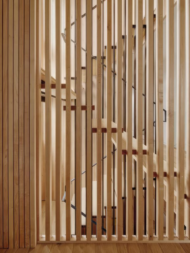 Interior architectural detail showing a tall, open timber stair enclosure or balustrade. Numerous slender vertical pale-wood battens frame and partially screen a stair rising through the space, with visible timber treads, landings, dark metal fasteners, and warm wood flooring. The composition emphasizes rhythm, transparency, craftsmanship, and vertical height; no furnishings are visible.