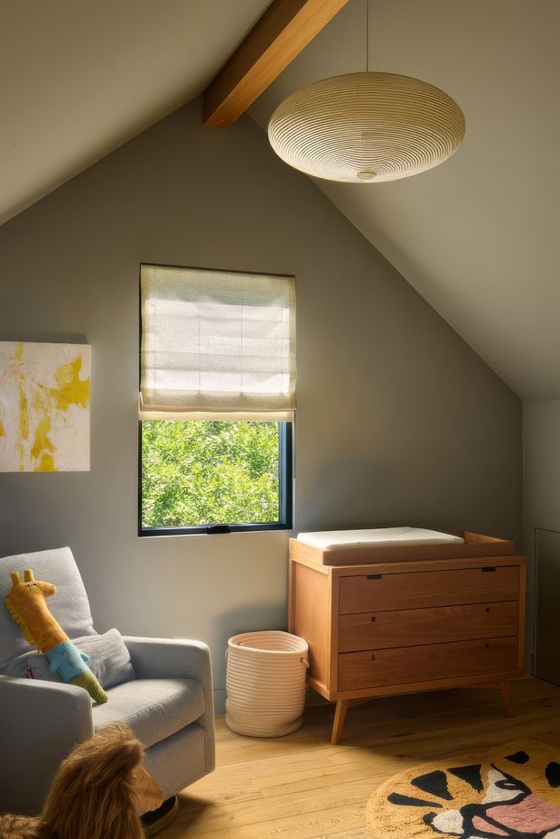 Warm, sunlit nursery or child's bedroom with a sloped ceiling and exposed timber beam, featuring a round woven pendant light, dark taupe walls, timber flooring, a mid-century-style wooden changing dresser, upholstered armchair, soft roller blind, and playful rug and toys. A window provides greenery views outside.