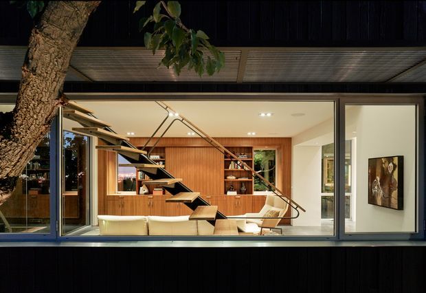 View from outside through expansive glazing into a refined contemporary living room. The dark-clad facade frames a warm timber-lined interior with built-in cabinetry, open floating stair treads, a light sofa, armchair, artwork, and large windows. A mature tree partially obscures the left side, while exterior canopy slats run across the top.