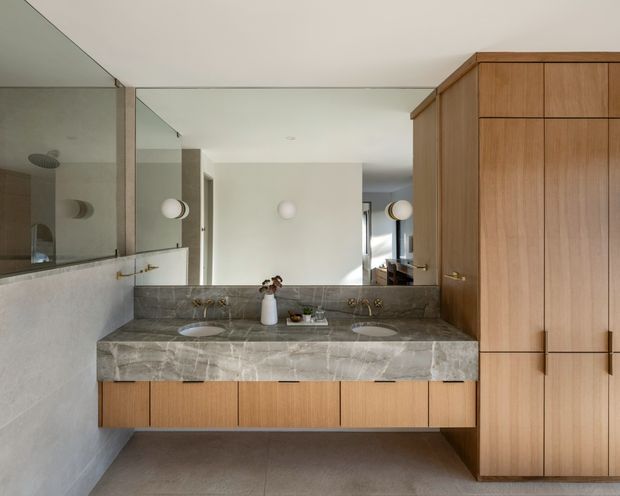 A refined bathroom vanity area with a wide double basin set into a grey veined stone benchtop, warm timber cabinetry, large wall mirror, pale plaster walls, and understated globe wall sconces. A glass-enclosed shower is visible at the left edge, while a full-height timber storage cabinet occupies the right side. The composition is clean, warm, and contemporary with a restrained material palette.