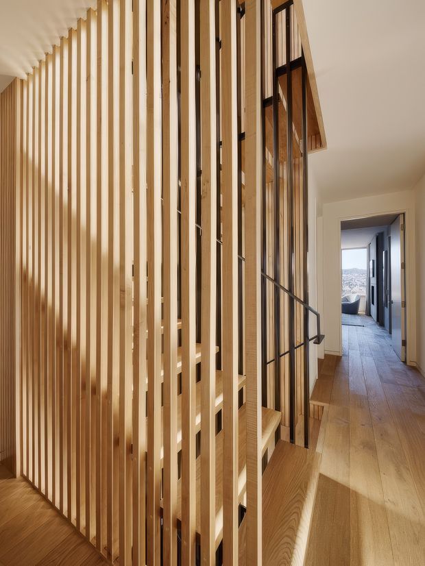 A warm, contemporary interior hallway featuring a sculptural timber slatted screen and stair balustrade. Vertical light-wood battens create a semi-transparent divider, with integrated timber steps and a dark metal handrail. The hallway continues toward a bright room with distant city views, and wide timber flooring runs throughout.
