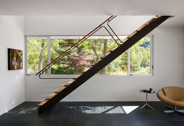 A minimalist interior showing a sculptural dark timber staircase crossing diagonally through the room, with slender metal balustrade detailing. Full-width windows overlook dense greenery, while a sculptural tan lounge chair and small side table sit on a dark tiled floor. White walls, strong natural light, and sparse furnishings create a refined contemporary atmosphere.