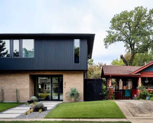 Exterior architectural photograph showing a contemporary two-storey home with a dark vertically ribbed upper facade, pale brick lower level, flat or low-pitched roofline, large black-framed windows and glazed entry doors. The house sits beside a colourful traditional timber bungalow with a pitched tiled roof. A landscaped front yard includes clipped lawn, gravel garden beds, architectural grasses, paved stepping stones and a concrete driveway.