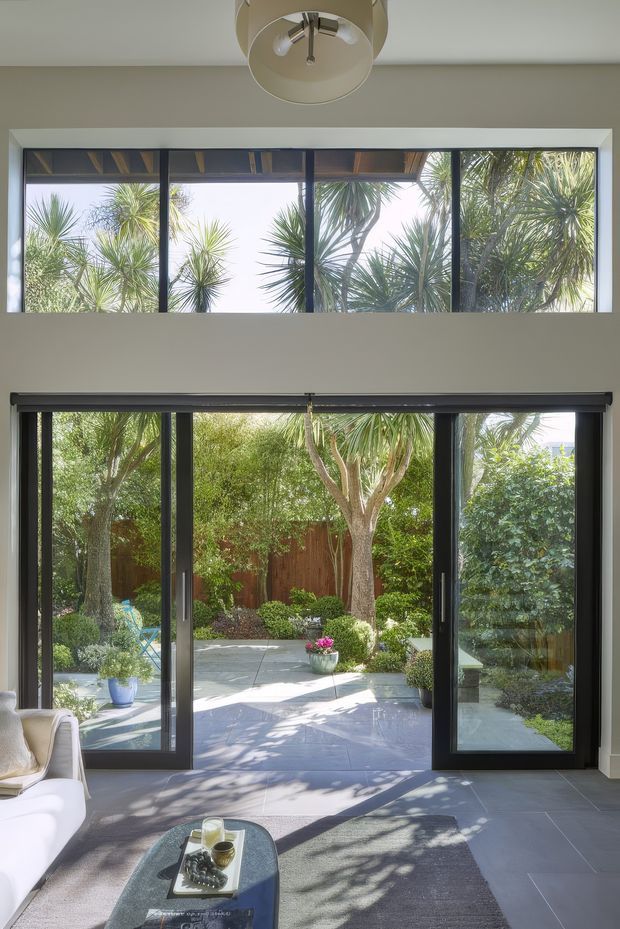Bright contemporary living room opening onto a landscaped courtyard through large dark-framed glazed bi-fold or sliding doors. A high horizontal clerestory window brings in additional daylight and frames dense sculptural garden planting. The room has a neutral plastered interior, grey tiled floor, area rug, pale sofa, low coffee table, and a simple ceiling light. Strong indoor-outdoor connection with abundant natural light and garden views.