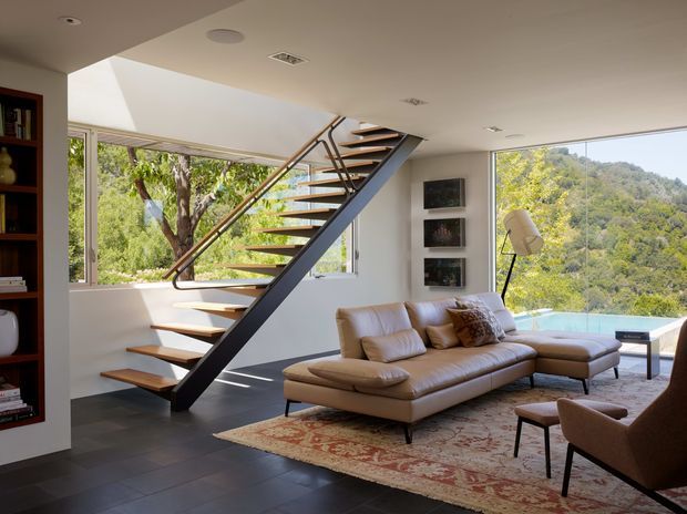 Bright contemporary living room with expansive windows overlooking a wooded hillside. A sculptural open-tread staircase with dark steel stringers and warm timber treads dominates the space, while a large tan sectional sofa, patterned area rug, armchairs, floor lamp, built-in shelving and framed wall art create a warm, refined seating area. Dark tiled flooring and a white ceiling complete the interior.
